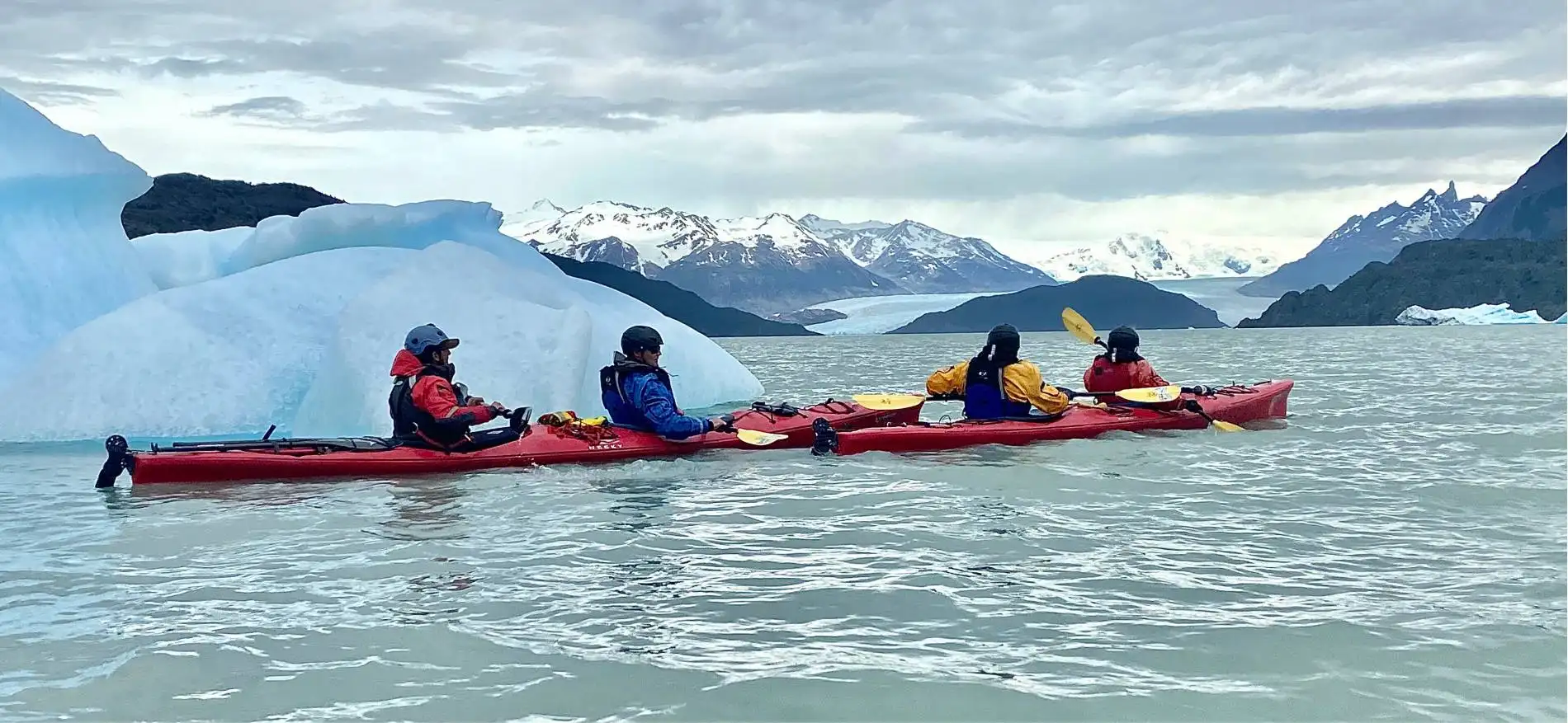 Dos kayaks dobles con cuatro turistas remando en las aguas del Lago Grey junto a un gran bloque de hielo flotante en la Patagonia.