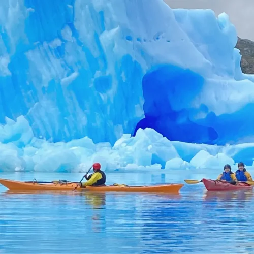 Two paddlers in a kayak in front of the vertical wall of a giant floating iceberg on Grey Lake.